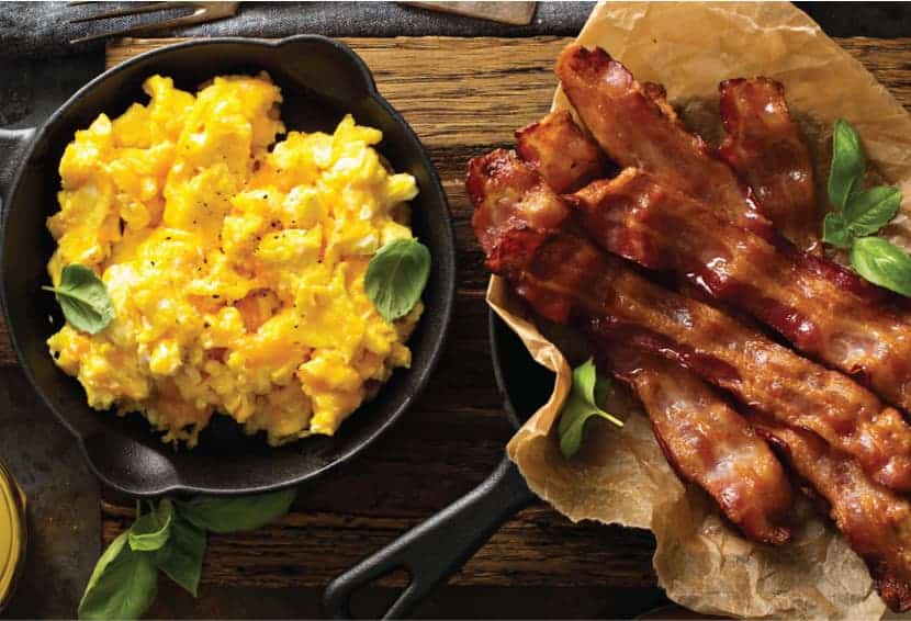 Scrambled eggs in cast iron skillet and another cast iron skillet with cooked bacon