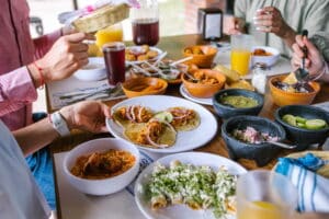 Top view of friends gathered around the table eating tacos. Focus is on the food on the table, only showing the peoples hands reaching for the different types of tacos and sides.