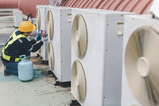 An Hvac Technician Wearing A Yellow Hard Hat And A High-Visibility Safety Vest Kneels On A Rooftop To Service A Row Of Industrial Air Conditioning Units. He Is Using A Manifold Gauge Set Connected To A Light Blue Refrigerant Tank To Check The System.