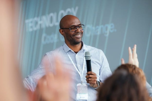 A Smiling, Bald Man With Glasses And A Striped Button-Down Shirt Stands On A Stage Holding A Microphone. He Is Wearing An Event Lanyard And Looking Toward An Audience Member Whose Hand Is Raised In The Foreground. In The Background, A Large Screen Displays The Words &Quot;Technology,&Quot; &Quot;Code,&Quot; And &Quot;Change.&Quot;