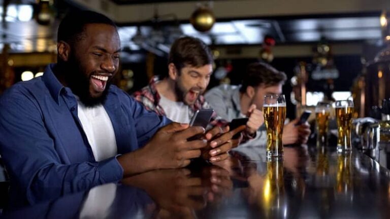 Three men sitting at a bar of a sportsbook while betting on their phones and drinking beer.