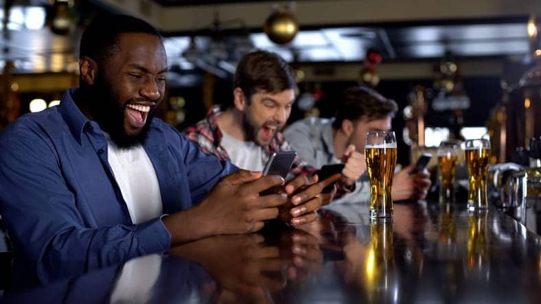 Three men sitting at a bar of a sportsbook while betting on their phones and drinking beer.