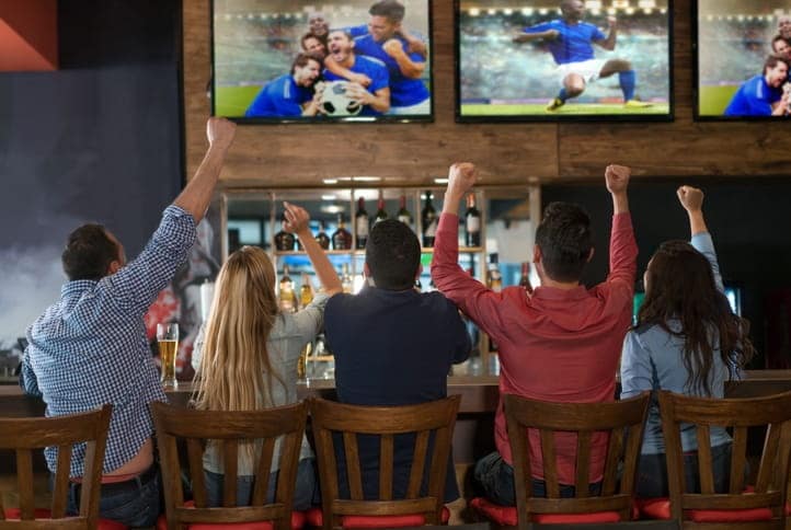 A row of five friends sitting at a bar, cheering for the soccer game on the television screens.