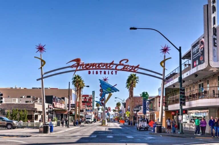 Iconic archway sign marking the entrance to the Fremont East District in Las Vegas, with retro neon decorations, palm trees, and a bustling street scene under a clear blue sky.