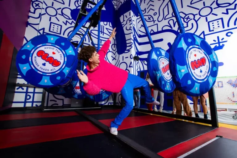 Person in a pink sweater and blue pants navigating oversized poker chip obstacles at PLAY Playground's Poker Parkour attraction, with bold graphic-art murals in the background