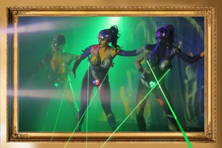 Three costumed performers with body paint and futuristic accessories wielding green laser beams on a fog-filled stage, framed within an ornate golden picture frame.